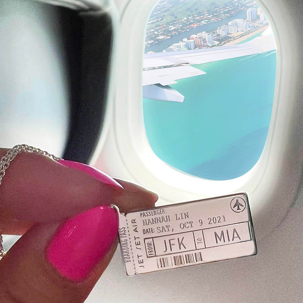 Photo of a Sterling Silver Boarding Pass Charm from the jewelry company Jet Set Candy. The charm is customizable and this one shows the name “Hannah Lin” engraved along with the airport codes “JFK” and “MIA” as well as a date. The photo was taken on a plane in front of an airplane window overlooking turquoise water and the Miami skyline. The charm looks very similar to a real boarding pass and includes an airplane icon and fake engraved bar code.