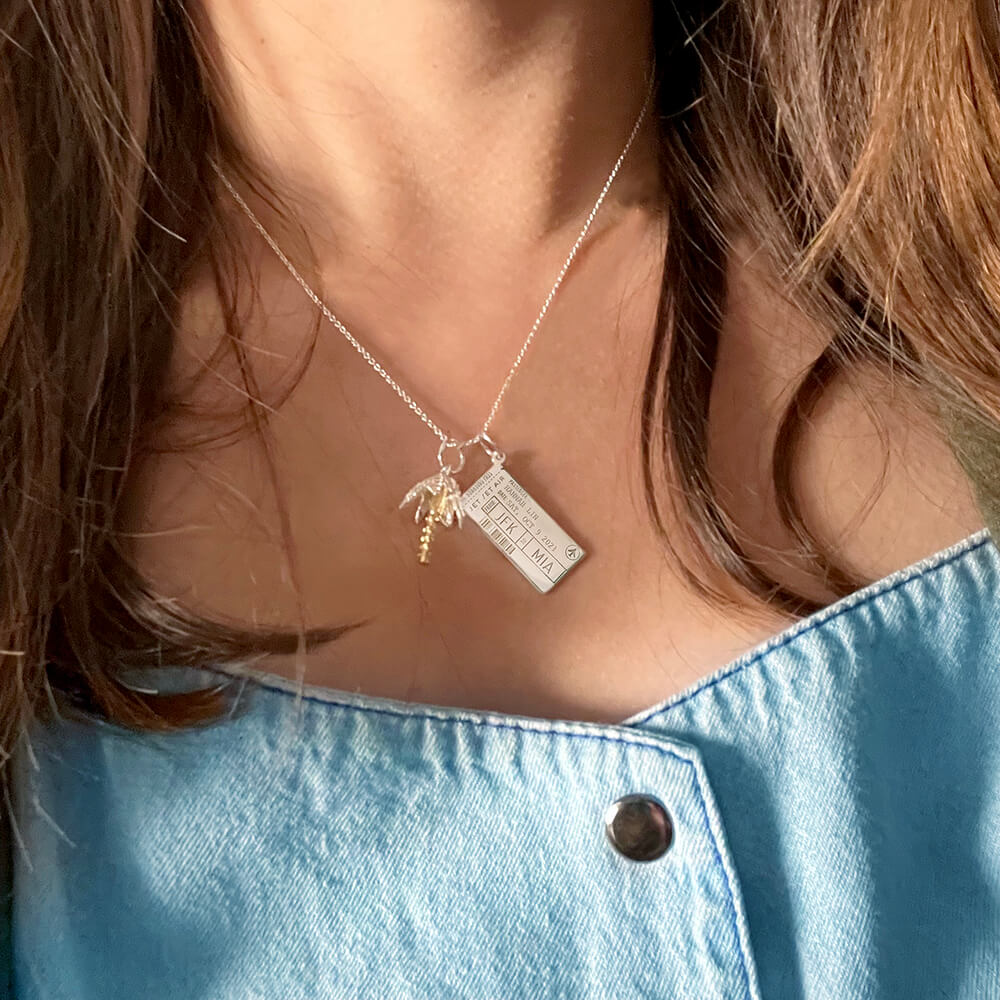 Photo of a woman with long brown hair wearing a charm necklace that includes a two-tone silver and gold palm tree charm as well as a Sterling Silver Boarding Pass Charm from the jewelry company Jet Set Candy. The boarding pass charm is customizable and this one shows the name “Hannah Lin” engraved along with the airport codes “JFK” and “MIA” as well as a date. The charm looks very similar to a real boarding pass and includes an airplane icon and fake engraved bar code.