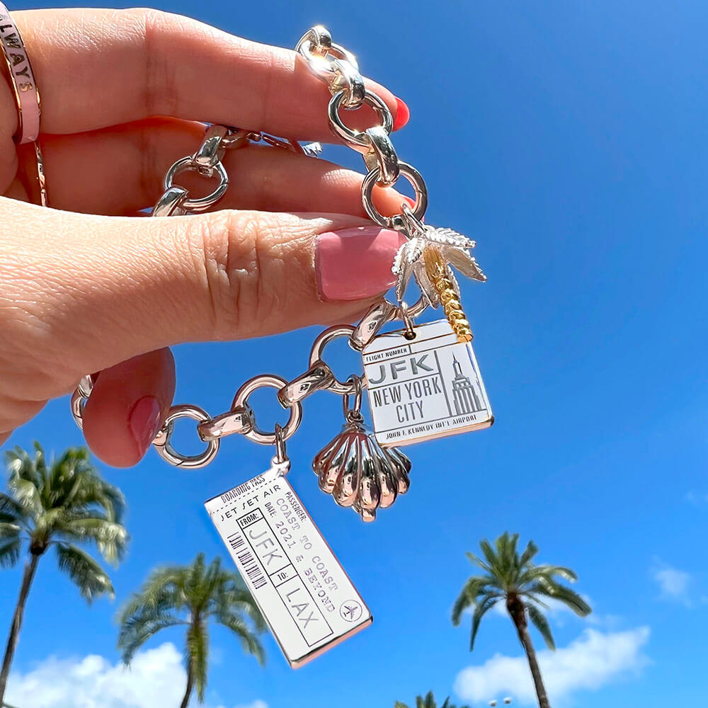 Photo of a hand holding up a Sterling Silver charm bracelet with a Boarding Pass Charm, a palm tree charm, a clamshell charm, and a New York City charm from the jewelry company Jet Set Candy.  The hand is in front of a blue sky with real palm trees in the background. The boarding pass charm is customizable and this one shows “LAX” and “JFK” as well as a date. 