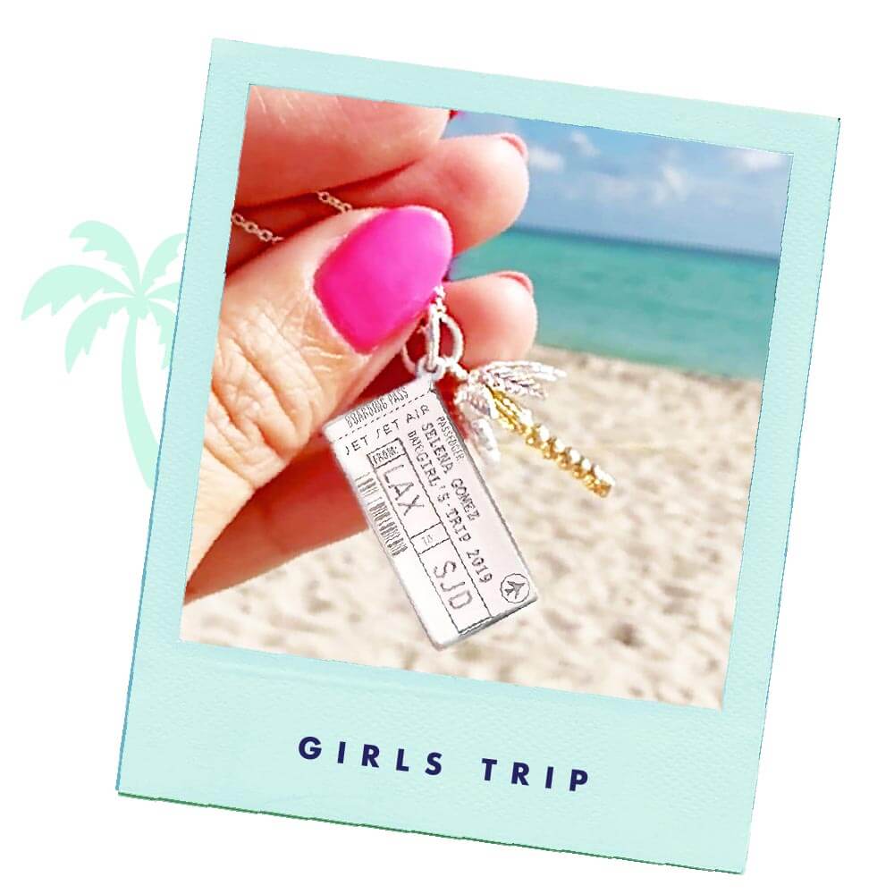 Photo of a Sterling Silver Boarding Pass Charm from the jewelry company Jet Set Candy. The charm is customizable and this one shows the name “Selena Gomez” engraved along with the airport codes “LAX” and “SJD” as well as a date. The photo was taken on a beach being held by a hand with hot pink nails. There is also a silver and gold palm tree charm being featured.