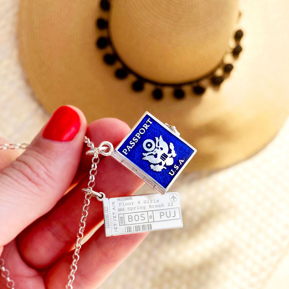 Photo of a hand holding up a Sterling Silver charm necklace  with a blue enamel and silver Passport Book Charm that opens as well as a Boarding Pass Charm from the jewelry company Jet Set Candy.  The hand is in front of a straw hat  The boarding pass charm is customizable and this one shows “BOS” and “PUJ” along with the owner’s name and a date.