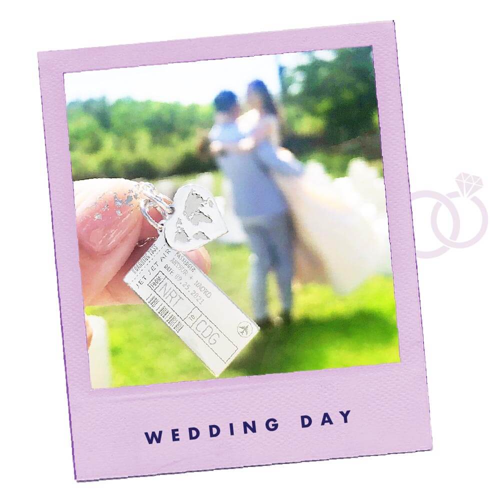 Photo of a purple polaroid frame with a close up of a hand holding a Sterling Silver heart charm with a world map engraved on it next to a sterling silver Boarding Pass Charm. The charms are held up in front of a couple who are getting married. The boarding pass charm from JET SET CANDY is engravable and this one features the couple’s names and the date and location of their wedding. On the polaroid frame are the words “WEDDING DAY”