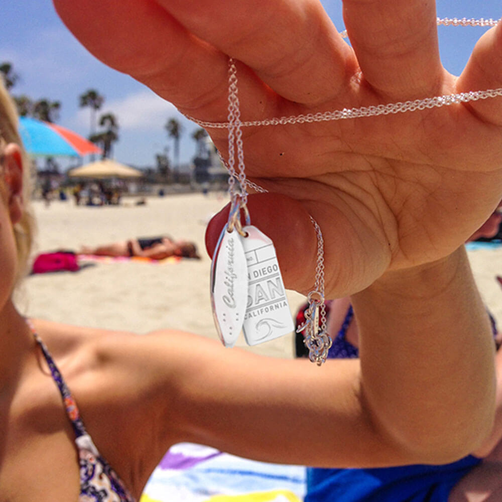 Woman at the beach holding up a sterling silver charm necklace featuring a 3D silver surfboard charm engraved with “California” and a silver SAN San Diego Luggage Tag souvenir charm.