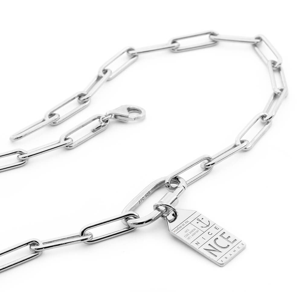 Photo featuring the Daily Charm Necklace in sterling silver from Jet Set Candy. The bracelet features long rectangular links with rounded corners and an inch-long carabiner that is shown with a silver travel charm for Nice France with the airport code NCE. The bracelet is shown snaked on a white background.