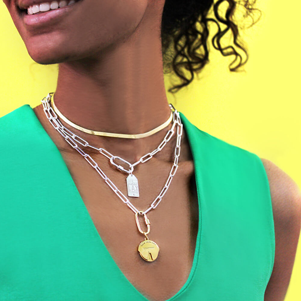 Photo of a model wearing a green top, a gold herringbone chain and two silver Daily Charm Necklaces from Jet Set Candy. The chain necklaces feature long rectangular links with rounded corners, each with an inch-long carabiner charm clip. One of the charm clips is holding a silver luggage tag charm and the other has a gold NYC spinner charm. The model is standing in front of a yellow background.
