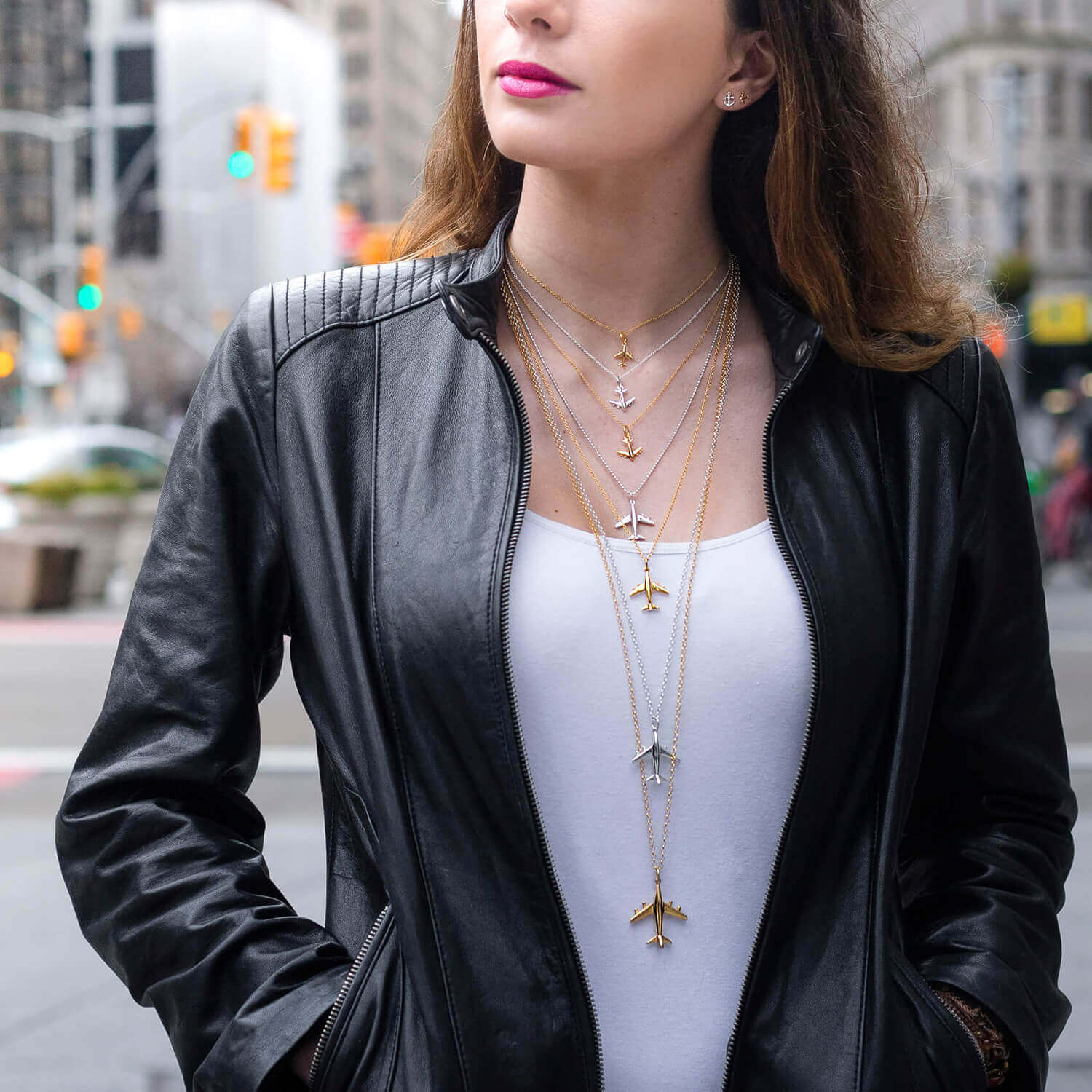 Image of a brunette woman wearing a white tank top and seven gold and silver necklaces in a variety of lengths. Each chain has an airplane charm hanging from it. The image is annotated in pink type with the varying lengths of the chains starting with the shortest shown at 14” and going all of the way to 36” long. The airplane charms come in a variety of sizes and the smallest ones are shown closest to her face and they get increasingly larger on the longer chains.