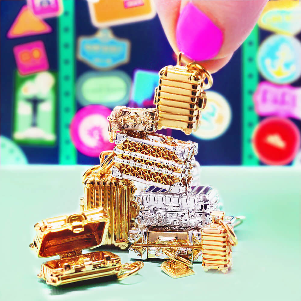 Image showing a stack of miniature sterling silver and gold suitcase charms stacked on top of each other with a hand picking the top one up. One of the charms looks like a vintage suitcase and the body of the charm is silver and there are gold travel stickers in different shapes that say “Paris” “India” and “NY”. Another has a pattern that resembles an iconic French steamer trunk. Another is a silver Rollie bag charm.