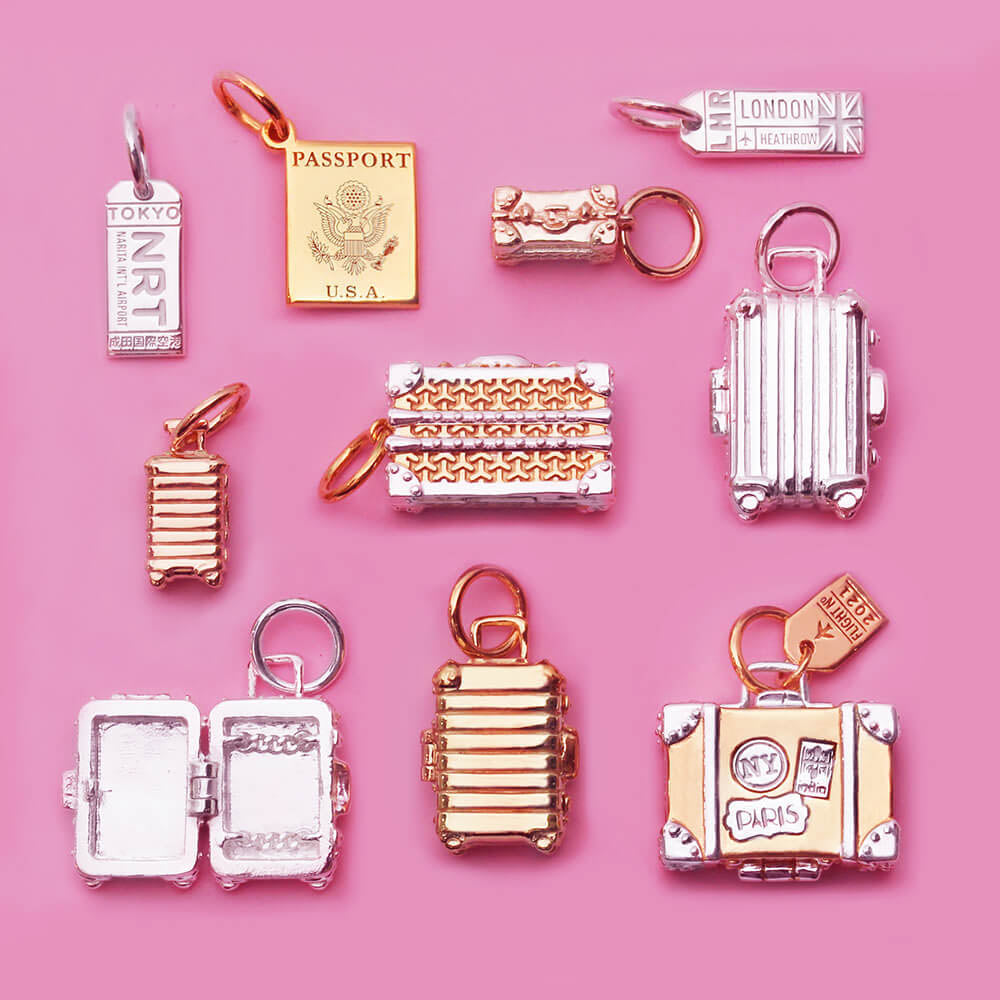 Image showing a variety of miniature sterling silver and gold travel charms on a pale pink background in a lay flat pattern.  One of the charms looks like a vintage suitcase and the body of the charm is silver and there are gold travel stickers in different shapes that say “Paris” “India” and “NY”. There is also a gold  passport book charm, silver luggage tag charms, and a variety of other suitcase charms, all made by Jet Set Candy.