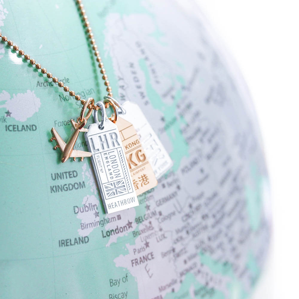 Travel charm necklace featuring a gold mini airplane charm, a silver London LHR Luggage Tag Charm, a gold HKG Hong Kong Luggage Tag Charm, and a silver JFK New York Luggage Tag Charm on a gold ball chain from Jet Set Candy. The necklace is shown on top of a mint green globe with map.