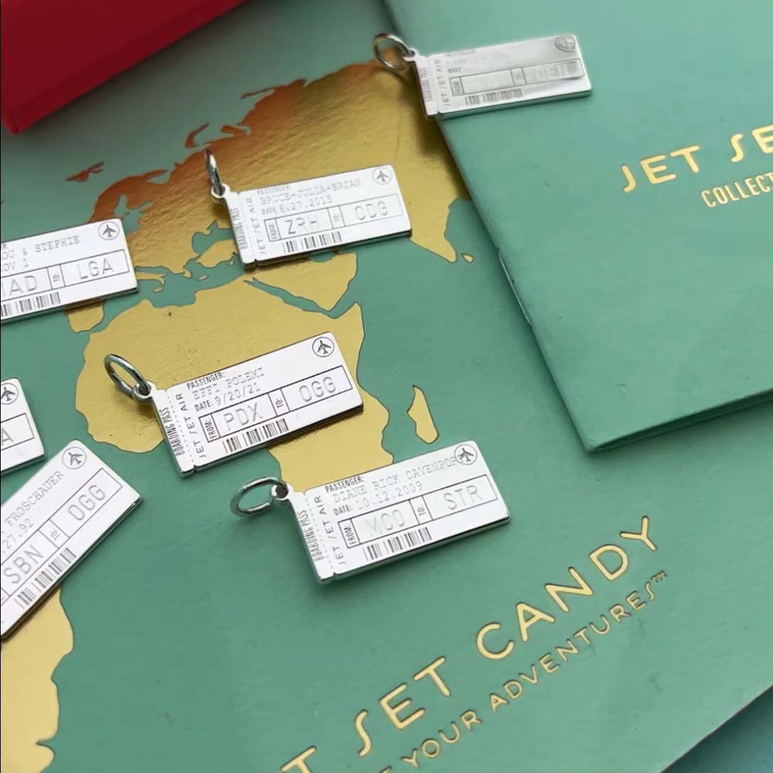 Photo of several Sterling Silver Boarding Pass Charms from JET SET CANDY shown on a mint and gold map background with the Jet Set Candy logo in gold. The boarding pass charms are customizable and each one shown here has different names, dates, and airport codes engraved on them. 