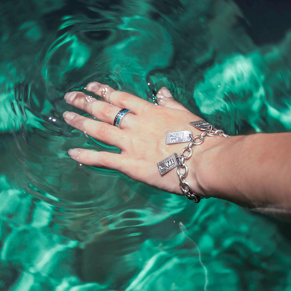Female arm with sterling silver travel-inspired charm  bracelet and charms for Tampa, Turks & Caicos, and Honolulu. The person  is also wearing a  black enamel and silver ring with the word WANDERLUST on it. The hand is touching a pool of deep green water. All jewelry is from Jet Set Candy